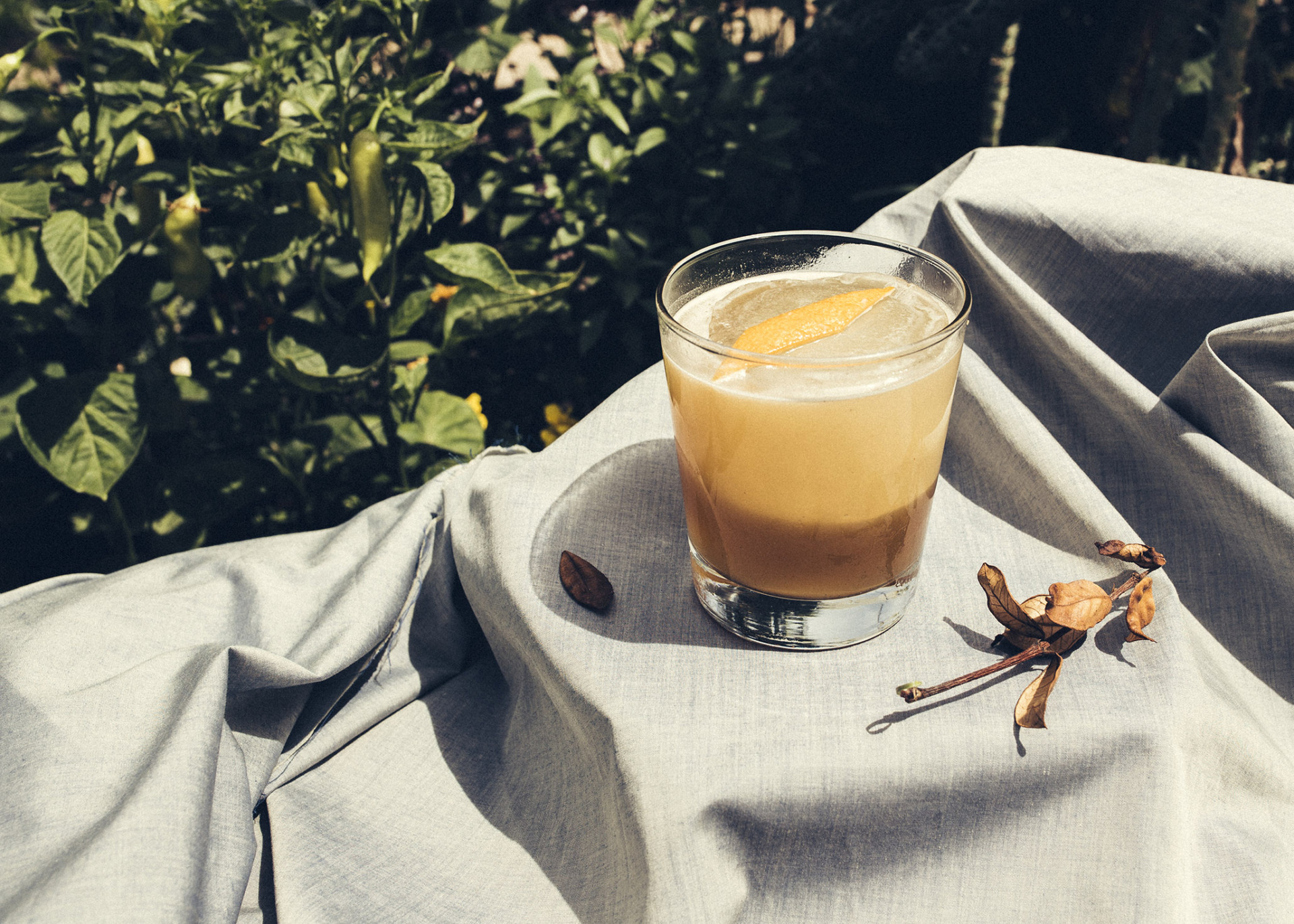 tan cocktail with orange rind on a gray tablecloth surrounded by a bush