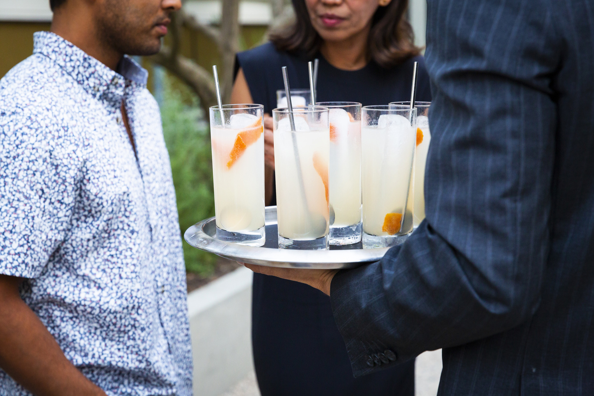 waiter in pinstrip suit holding out tray filled with white and orange cocktails for guests