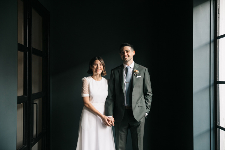 bride and groom holding hands in a dark green hallway by large window