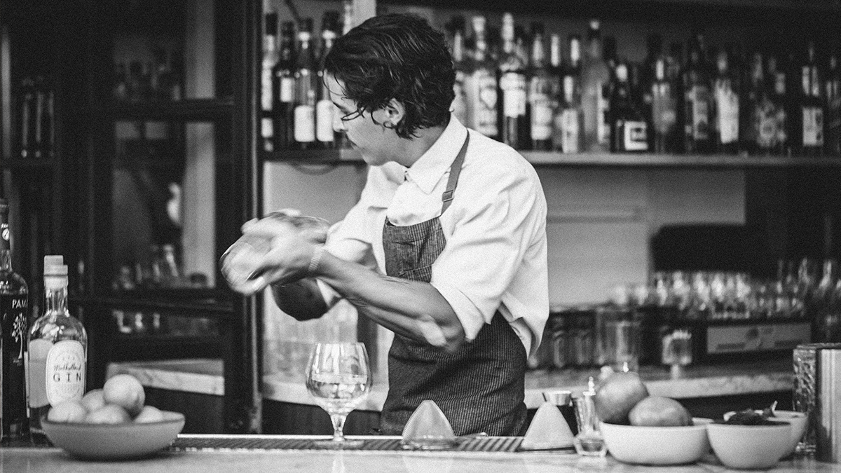 Bartender in black and white shaking cocktail behind bar.