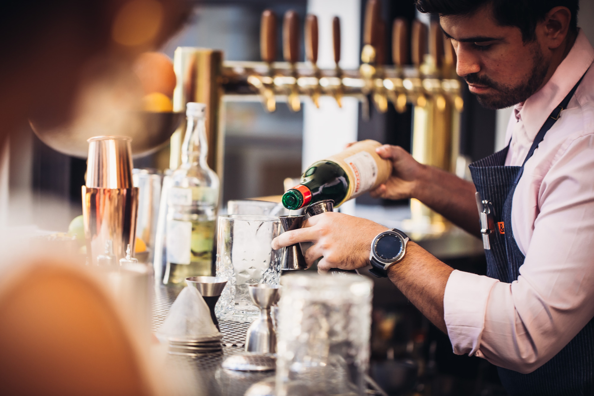 Bartender pouring spirit into a mixer behind a bar. Bartender is wearing a blue apron, pink shirt and has a big watch.