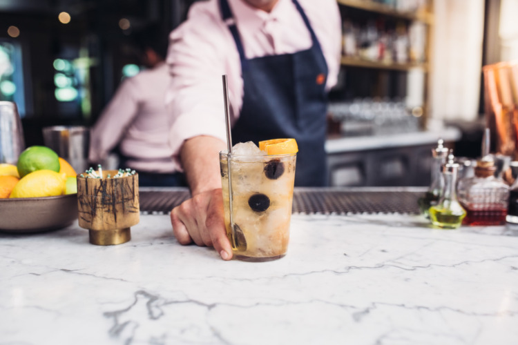 Cocktail being presented on a bar by bartender in blue apron and pink shirt, cocktail has frozen red grapes and orange on top