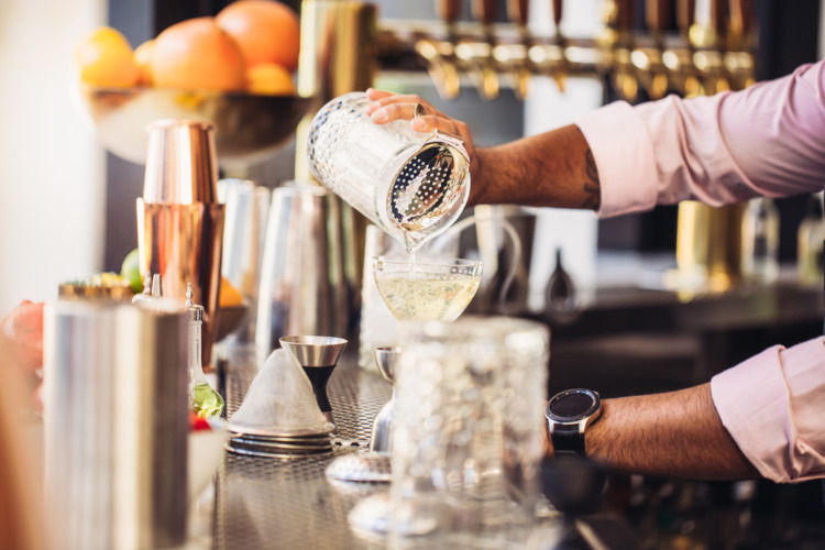 Male hands pouring yellowish cocktail out of a shaker into a pretty glass