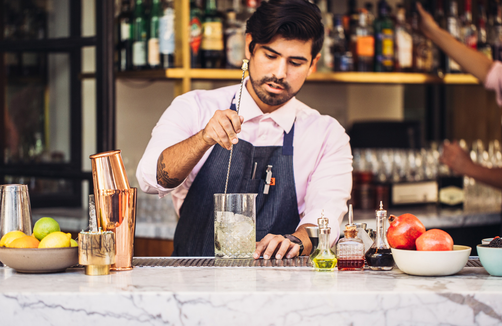 Male bartender with beard, pink shirt, blue apron stirring a yellowish cocktail