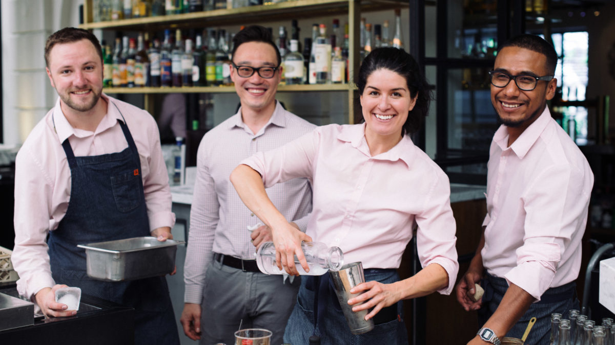 Four people wearing pink shirts smiling at a bar with bottles in background