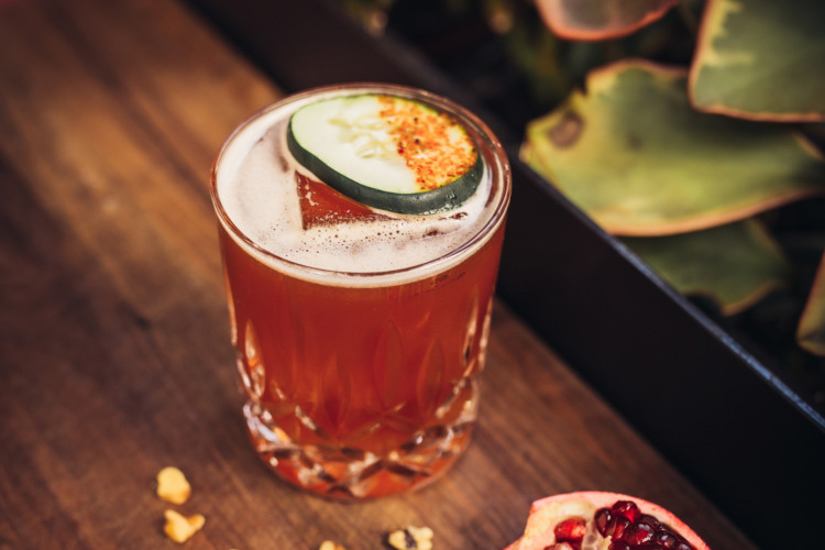 cocktail with cucumber and spices on top of a wood table with plants and pomegranate next to