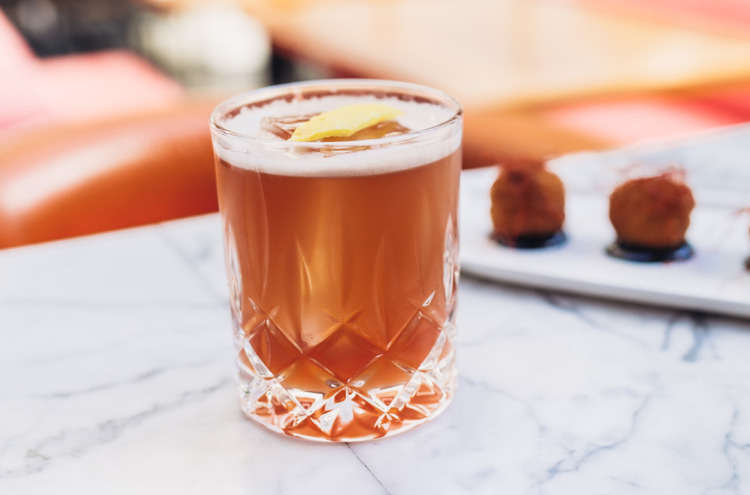 brownish cocktail with lemon twist sitting on a marble bar