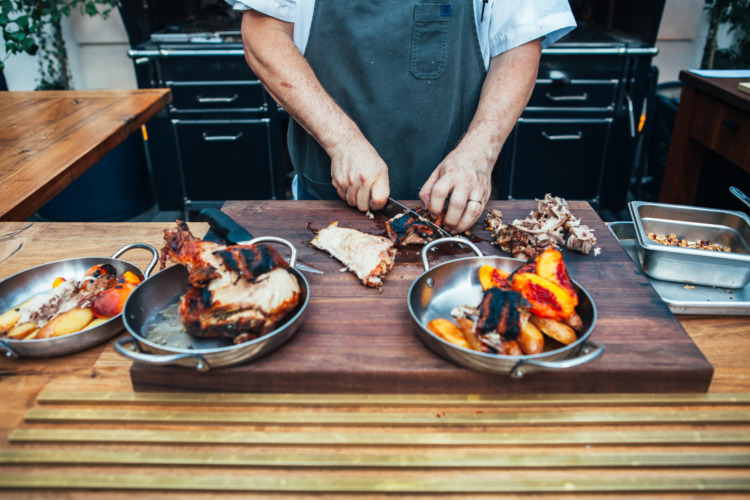 man cutting chicken and grilled peaches on table in front of a grill