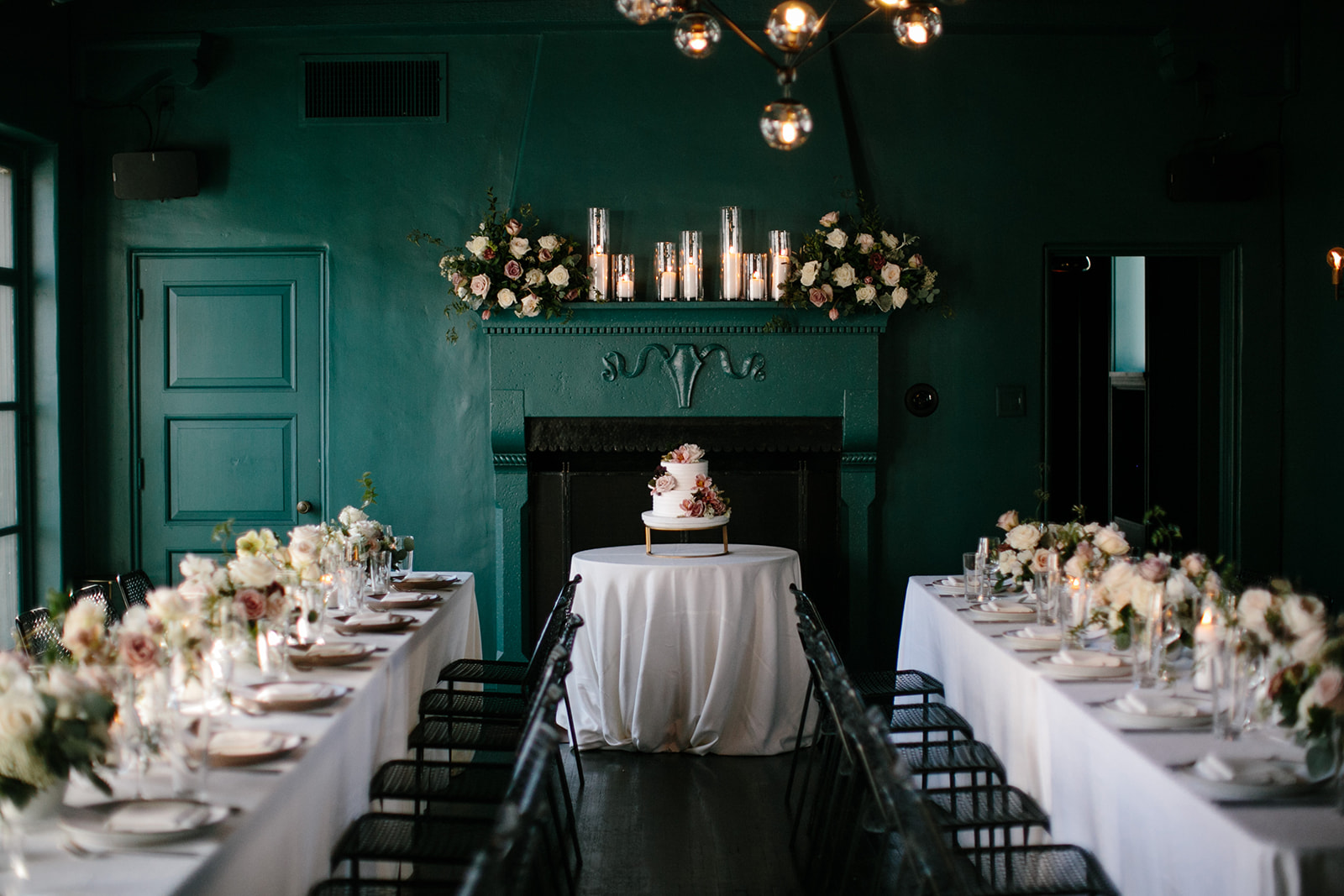 a wedding reception setup with dining tables and a cake table in front of a fireplace in a teal room. Cardinal's Quarters at Redbird in DTLA