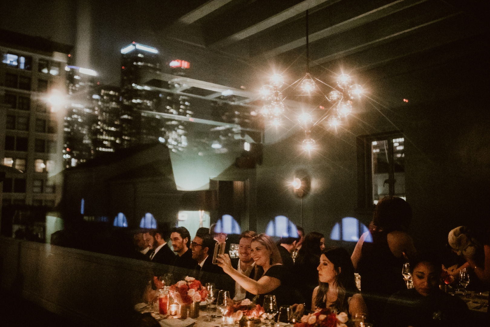 wedding guests sit at a wedding reception table, the downtown LA skyline is reflected in the photo, overlaid with the guests. Cardinal's Quarters in Downtown LA at Redbird