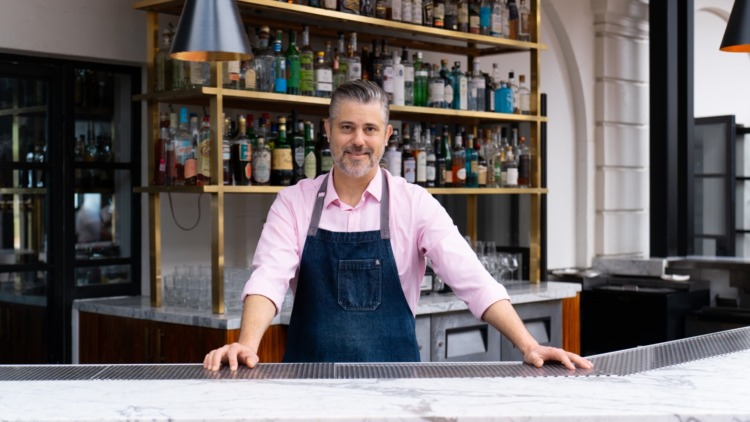 Tobin Shea, a tall man with greying hair and facial hair, standing at the redbird bar in a pink shirt and denim apron