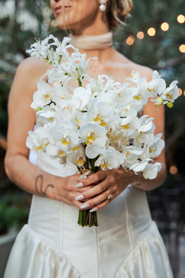 a bride holds her bouquet of white flowers with trees and string lights in the background