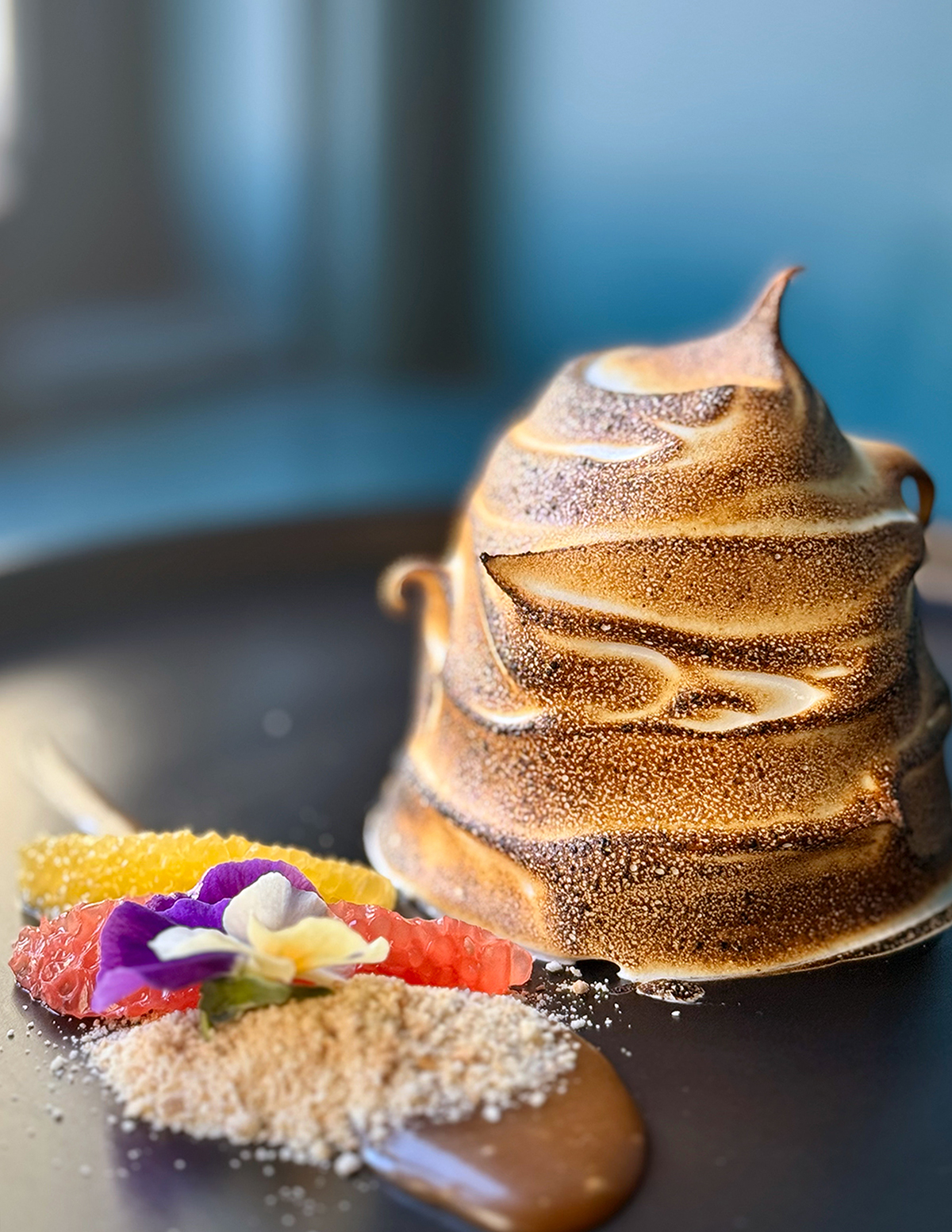 baked alaska with torched meringue sitting on a dark plate next to colorful garnish with teal walls in the background