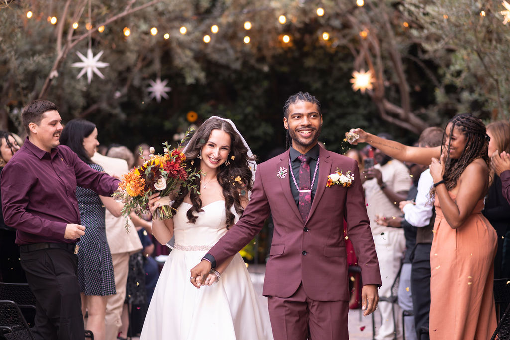 a bride and groom walk down the aisle at their wedding. the bride is wearing white holding a bouquet of flowers, the groom is holding her hand in a burgundy suit