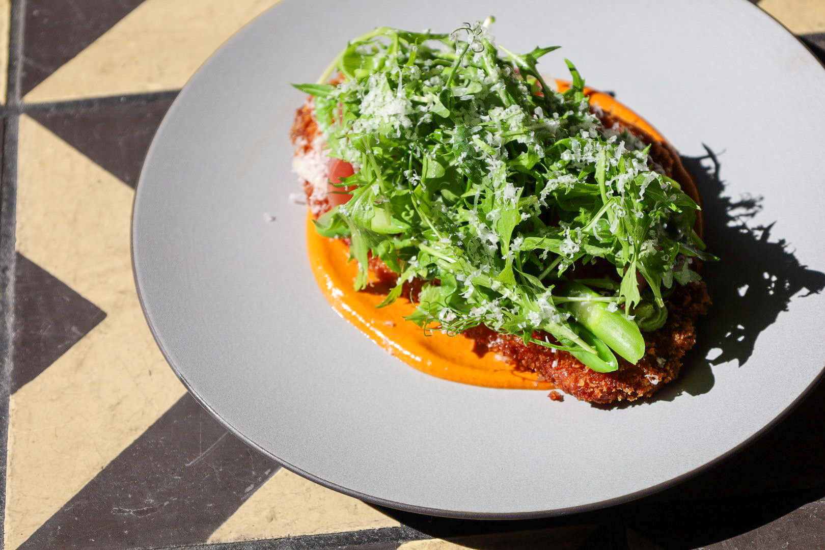 Green arugula on top of a fried chicken cutlet, on top of an orange sauce on a grey plate.