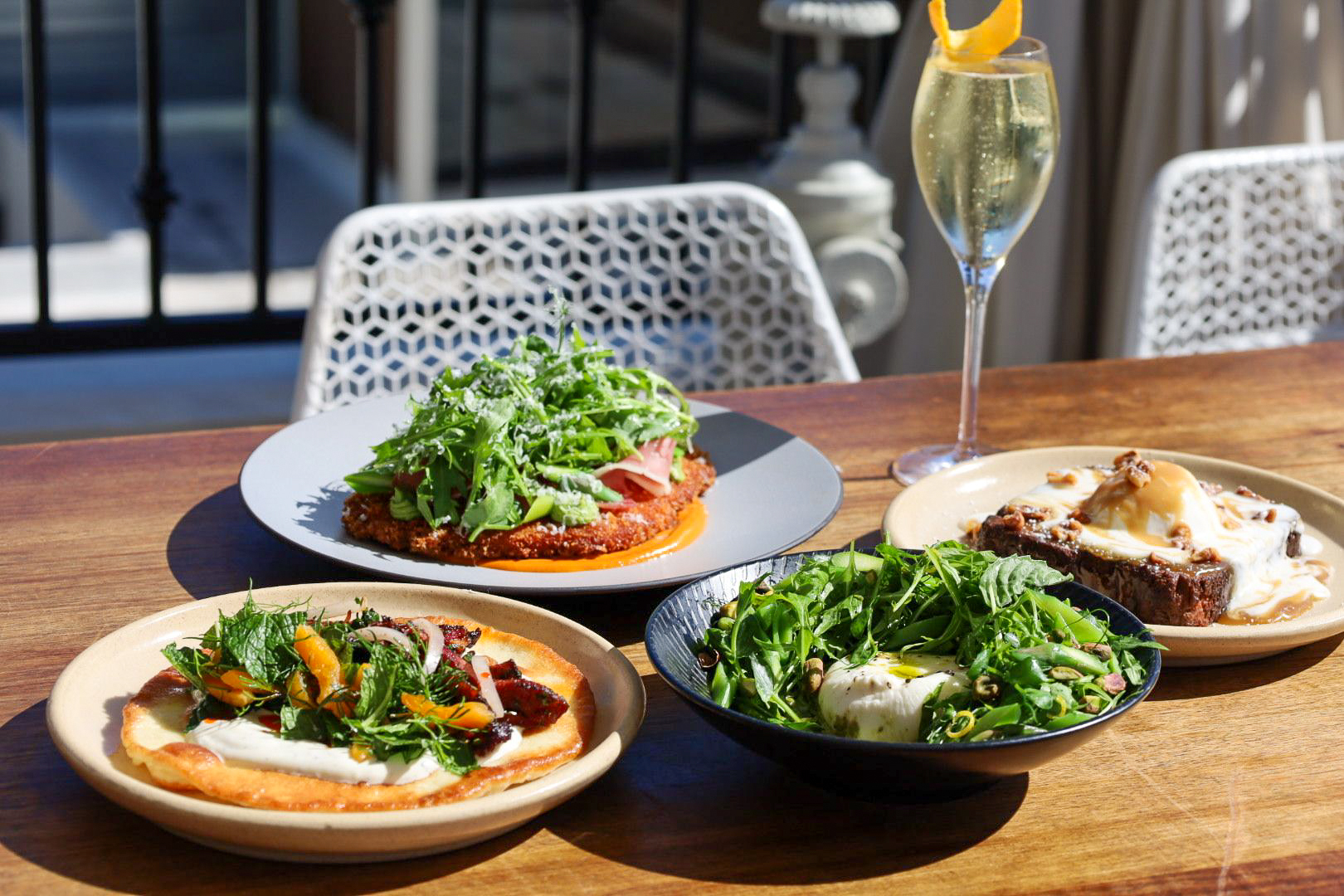 four dishes and a cocktail as an Easter brunch spread. Yellow flatbread with yogurt sauce and meat, breaded chicken with green arugula on top, a green salad with white burrata cheese in the middle, a french toast with ice cream on top, and a bubbly drink in a flute with a an orange twist on the rim.