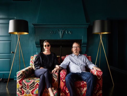photo of Amy and neal fraser on a colorful couch with teal walls wearing glasses