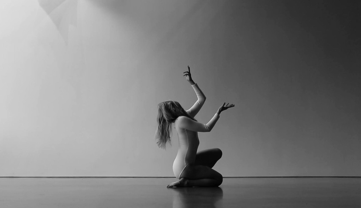 black and white artistic shot of a woman in a leotard posing on the ground with her hands raised