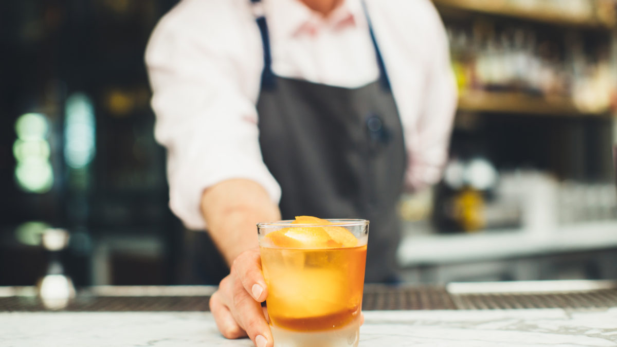 bartender holding out a cocktail with large square ice cube and orange twist