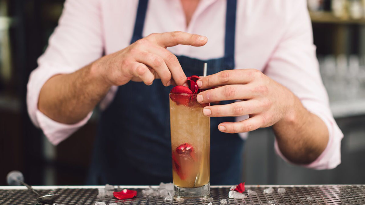 A bartender putting red rose petals into a tall cocktail glass