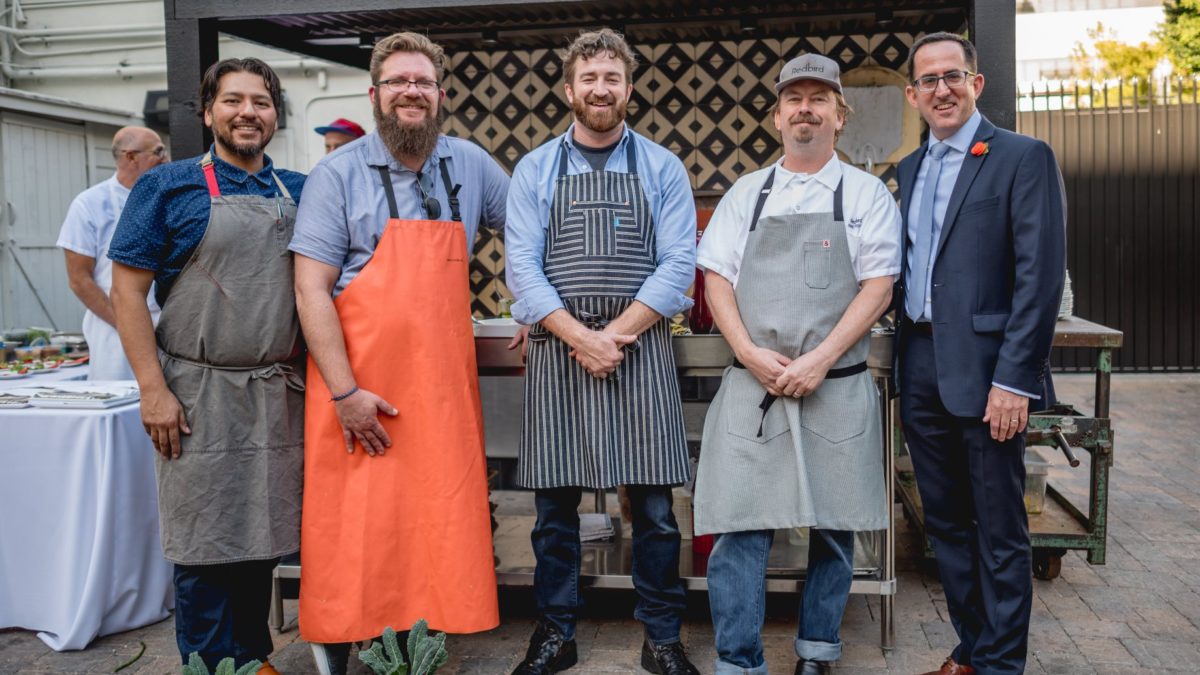 5 men standing in front of an outdoor food setup while wearing aprons