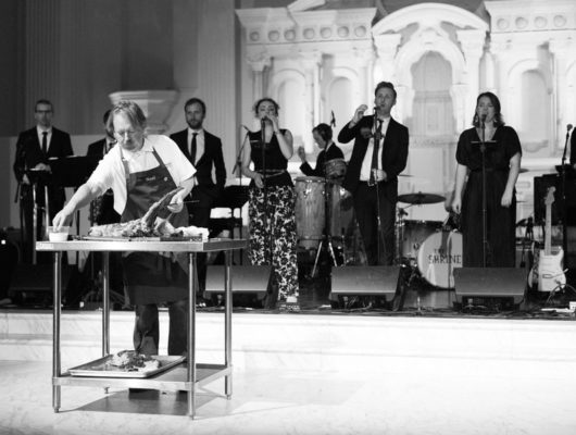 black and white photo fo group of people singing on stage behind a chef with tray of meat