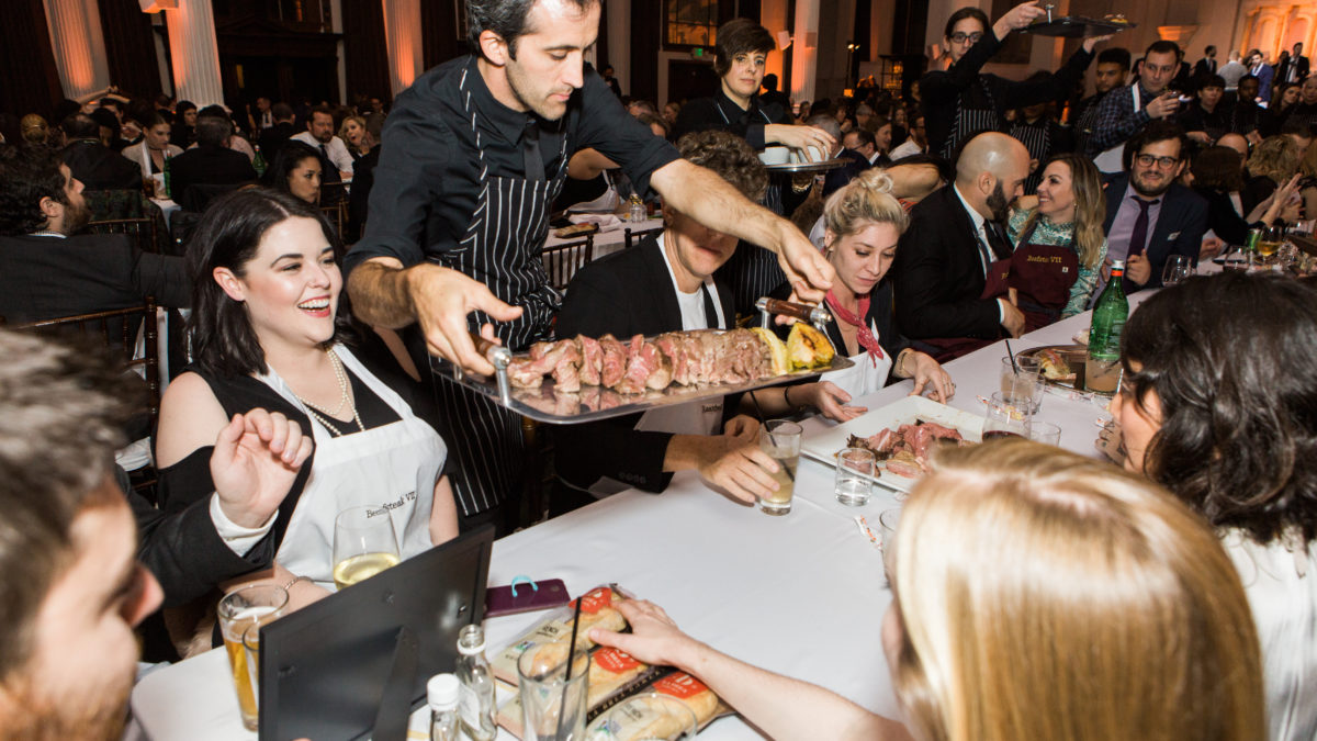 waiter putting out a tray of meat on a long table filled with people