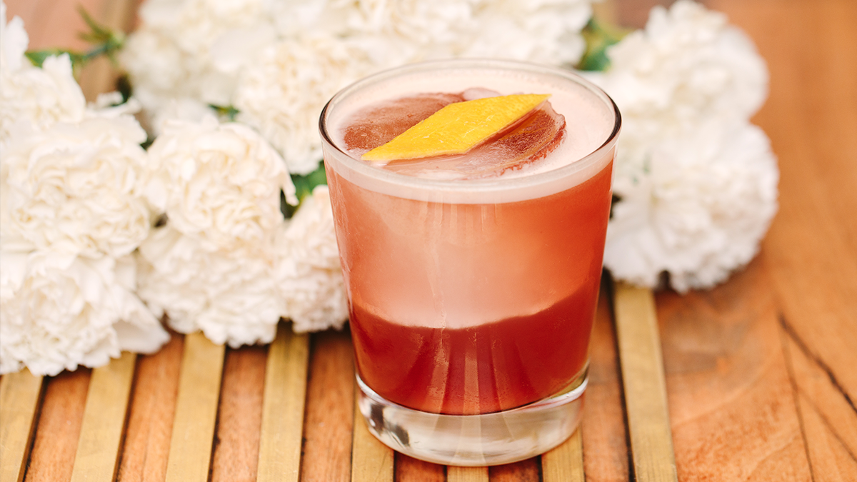 red cocktail with square ice cube and lemon rind surrounded by white flowers