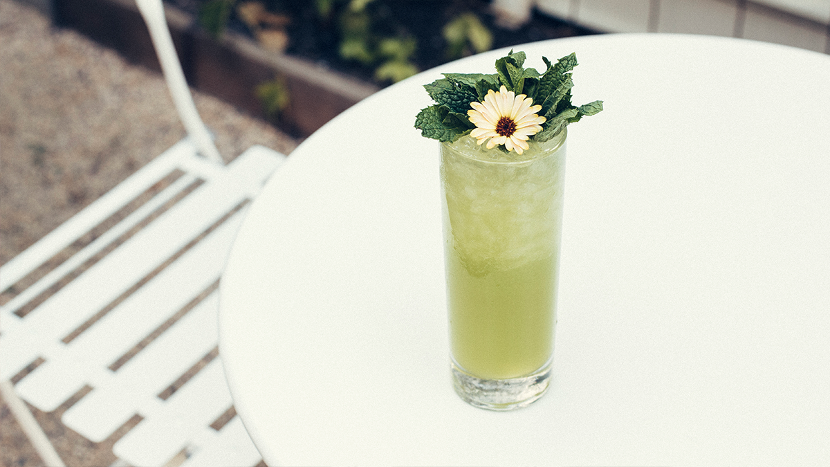green cocktail topped with daisy and mint on an outside white table