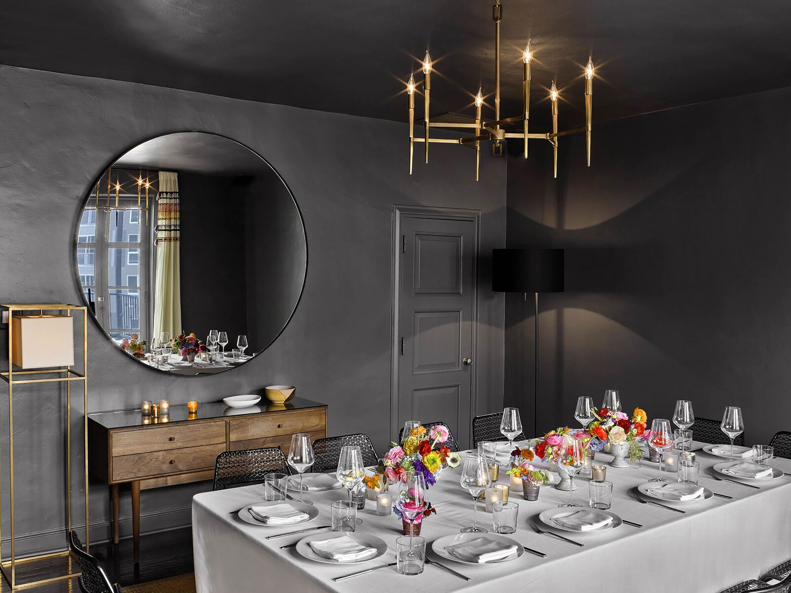 black room with gold light fixture, large round mirror, and large table set with flowers