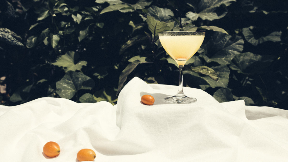 cocktail glass on outdoor table surrounded by three small oranges