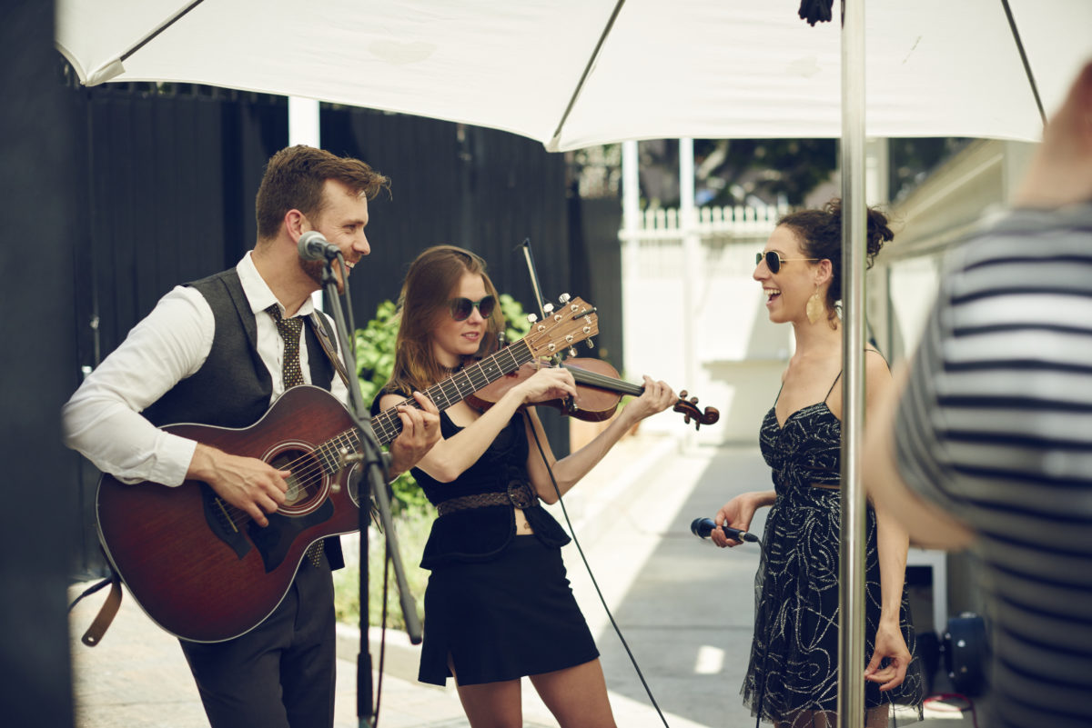 outdoor patio with man playing guitar, woman playing fiddle, and another woman holding a microphone
