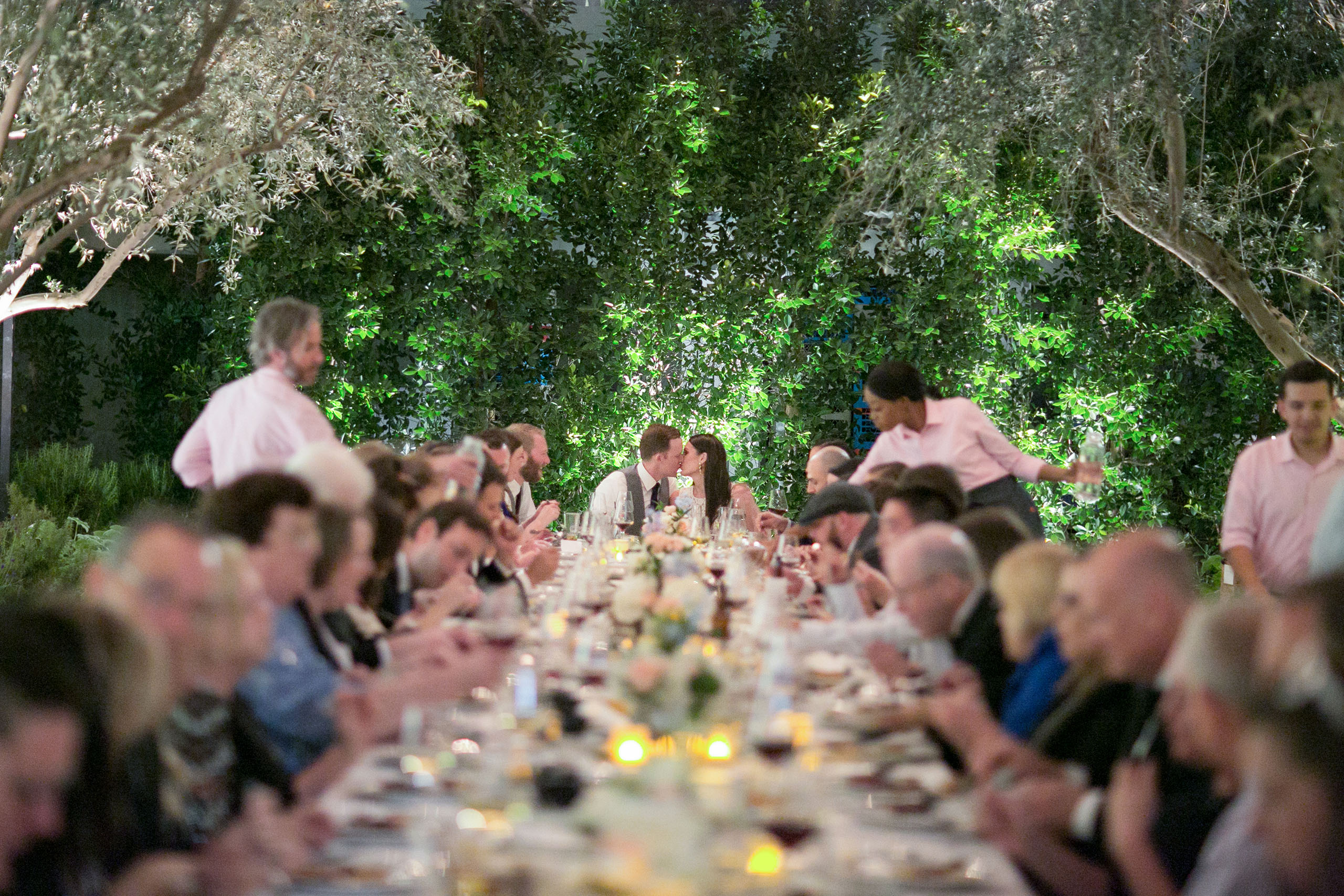 long table outside surrounded by trees with bride and groom kissing at end