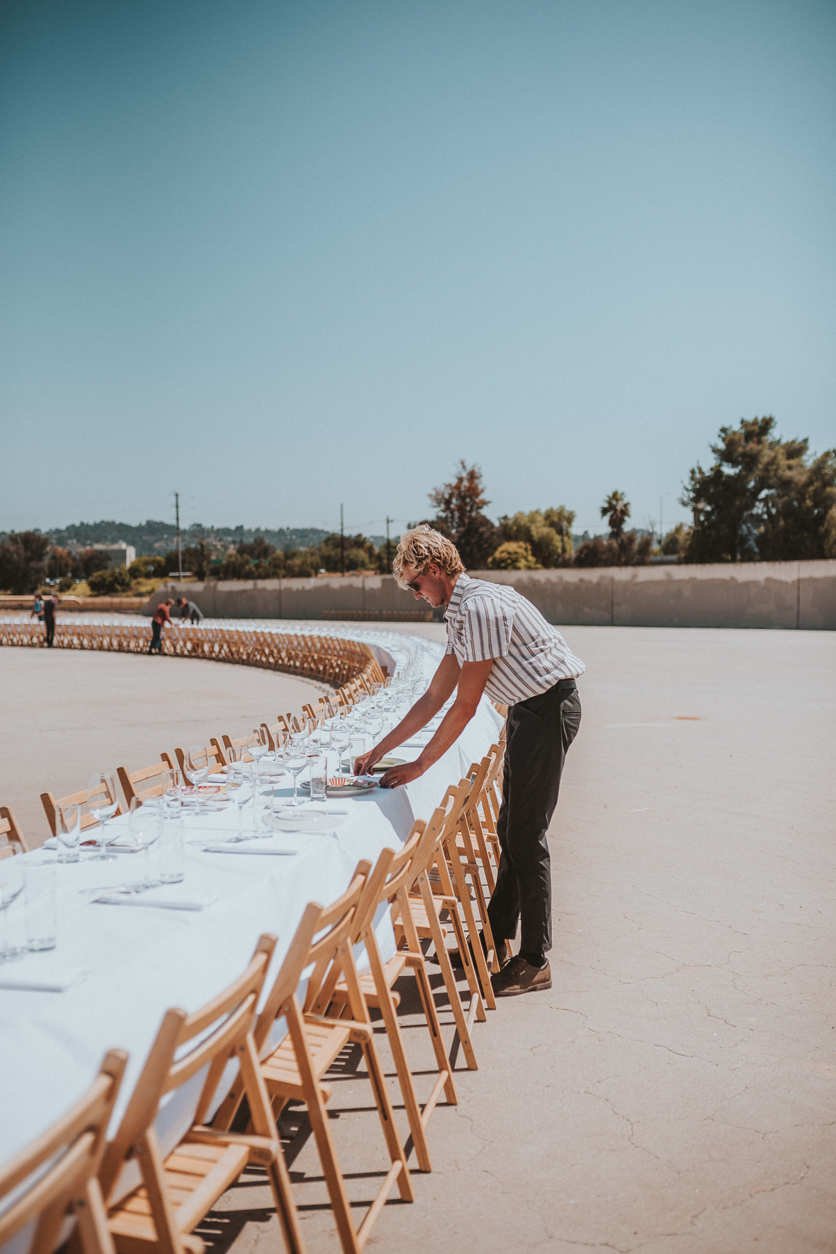 person in stripped shirt and black pants setting up place settings at a very long curved table outside