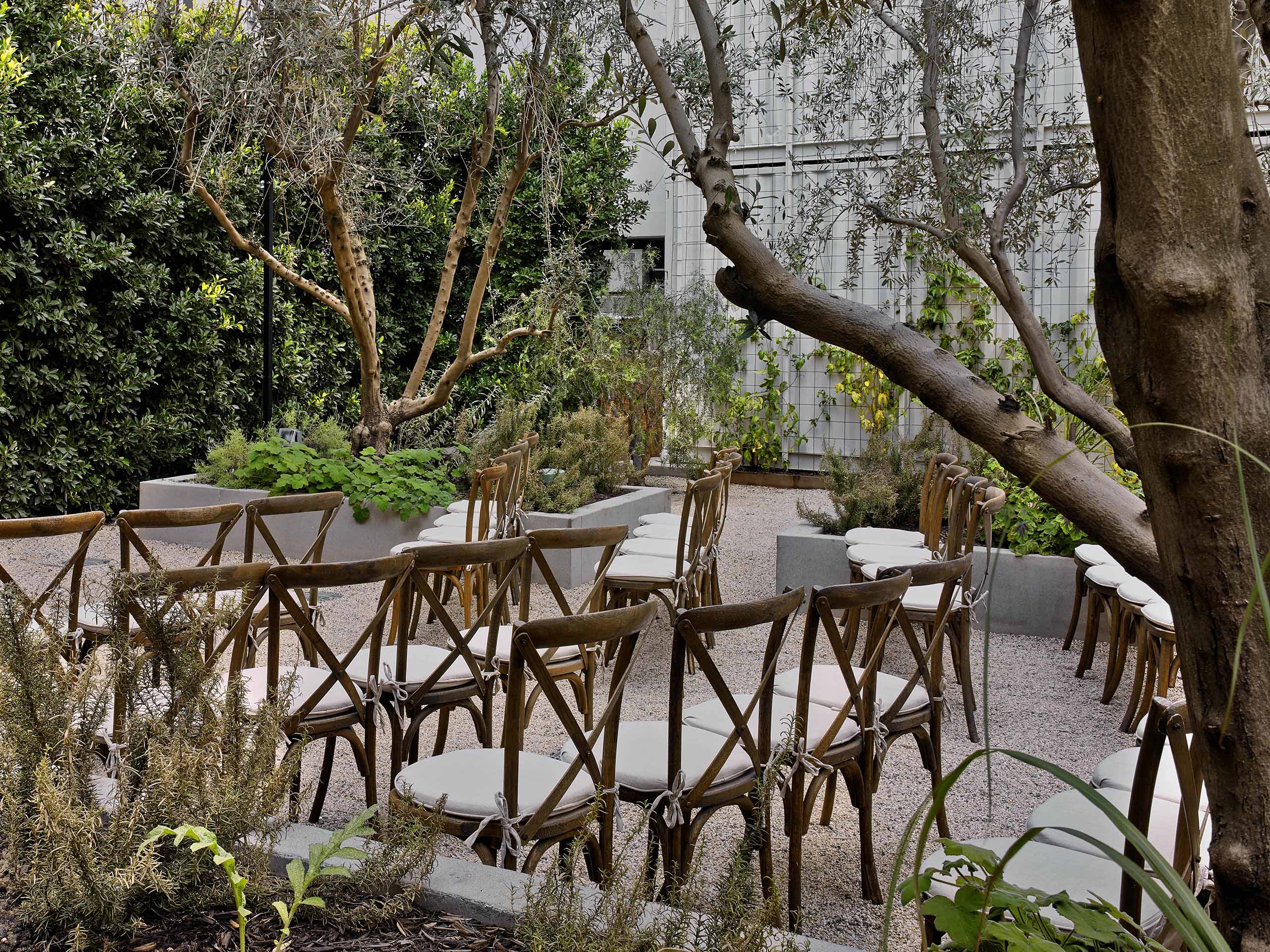 outdoor wedding setup in garden with two aisles of wooden chairs with white cushions