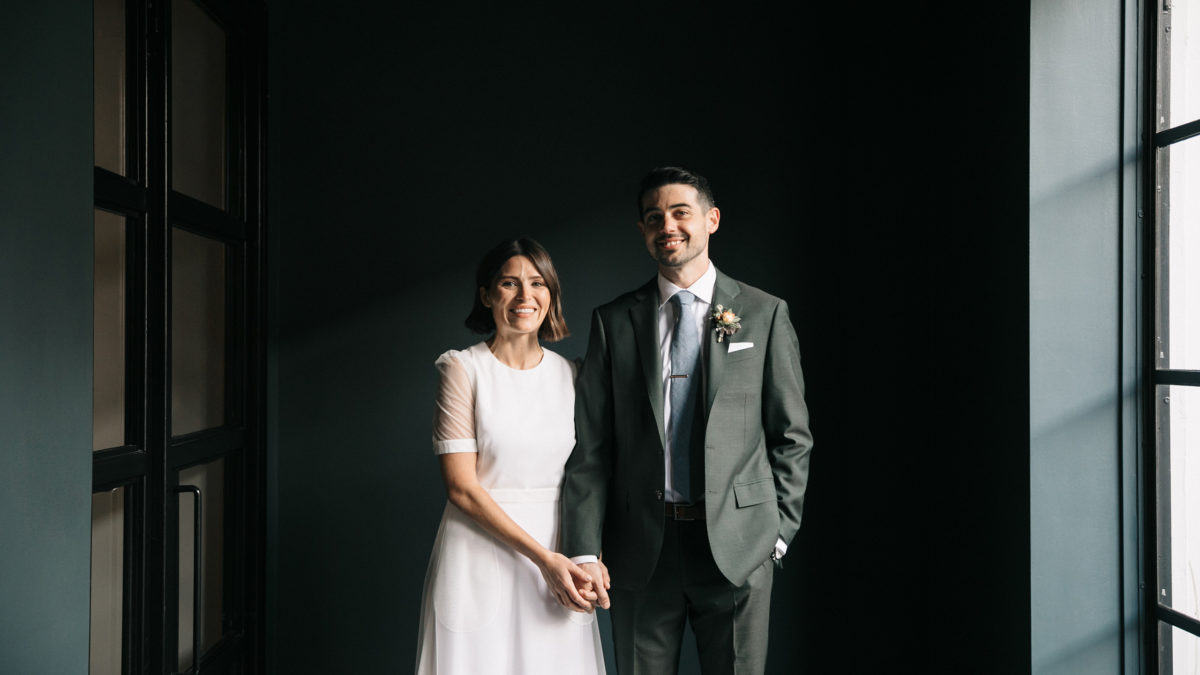 bride and groom holding hands in a dark green hallway by large window