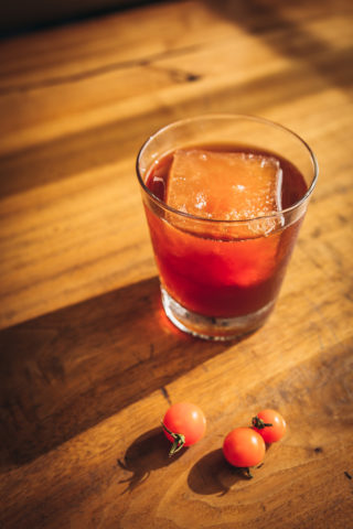 Red cocktail with big rock in a rocks glass with three small tomatoes in front of the glass.