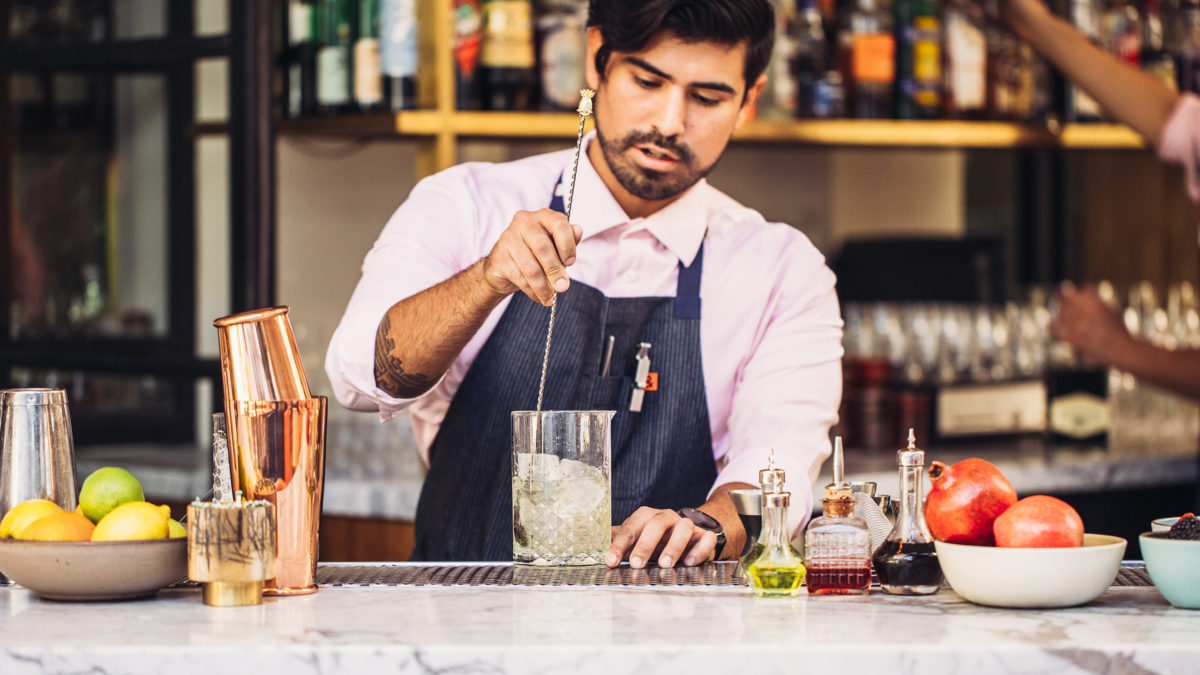 Male bartender with beard, pink shirt, blue apron stirring a yellowish cocktail