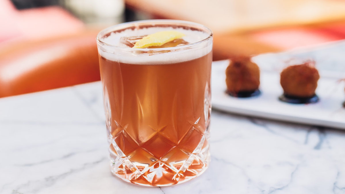 brownish cocktail with lemon twist sitting on a marble bar