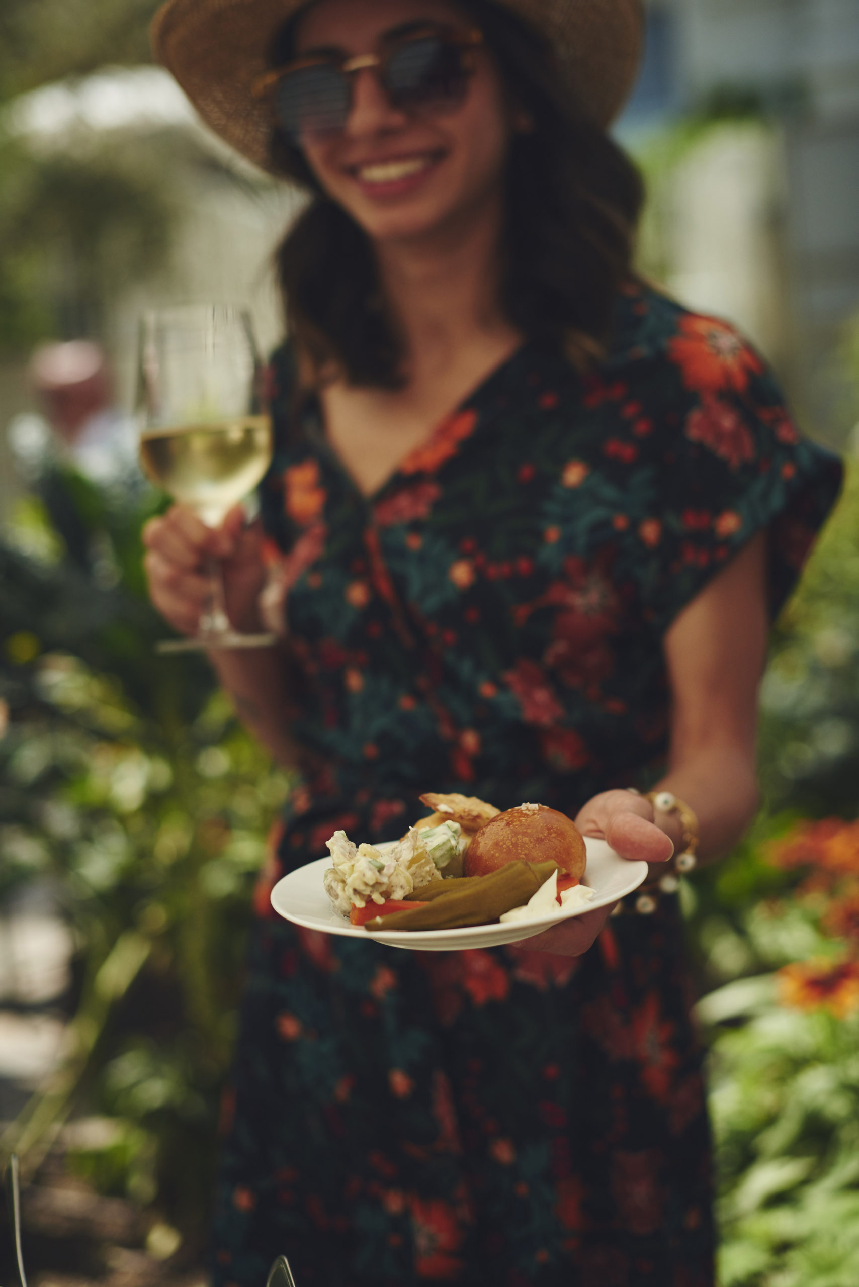 girl holding plate of food and glass of wine in colorful flower dress in a garden