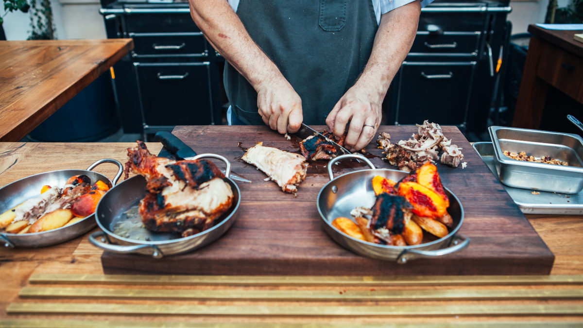 man cutting chicken and grilled peaches on table in front of a grill