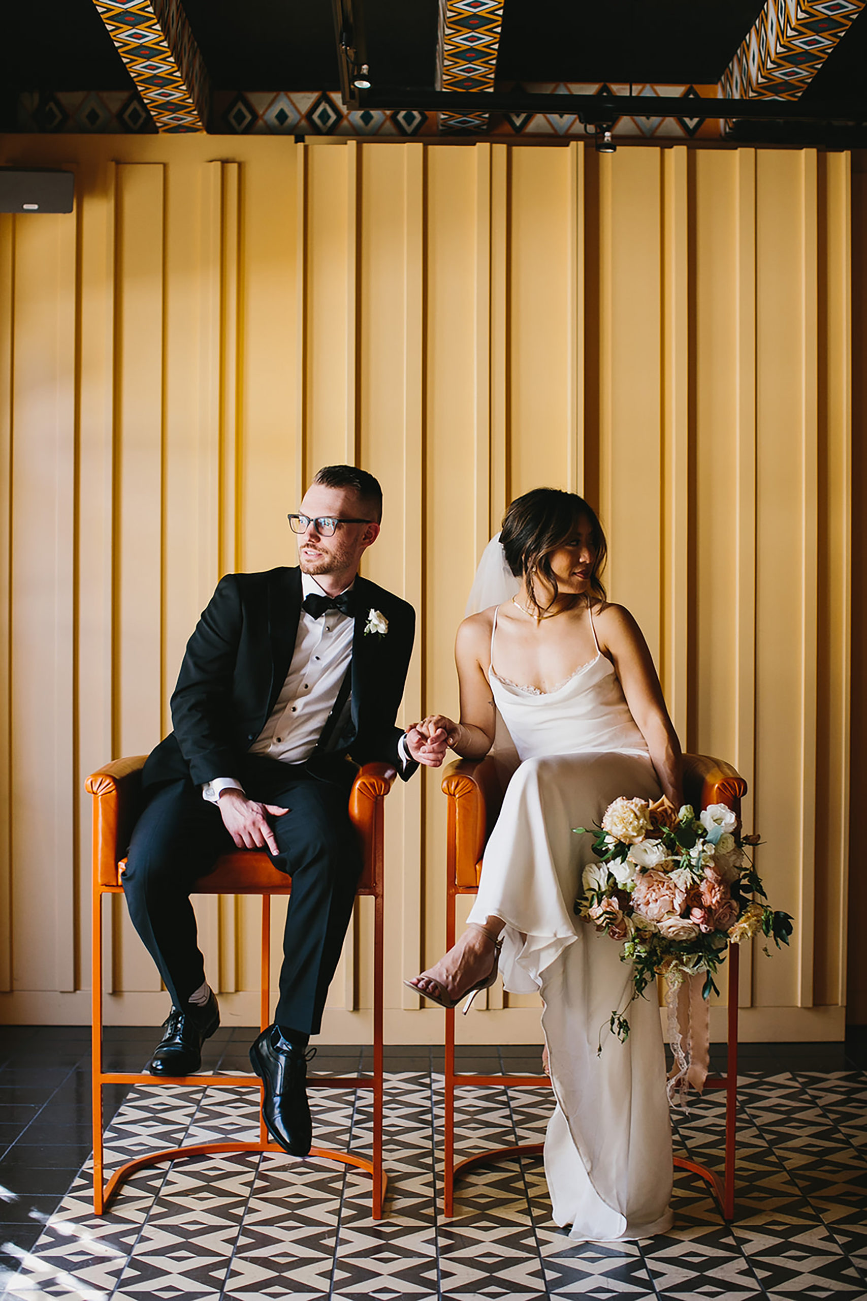 bride and groom holding hands siting in orange chairs behind a yellow wall