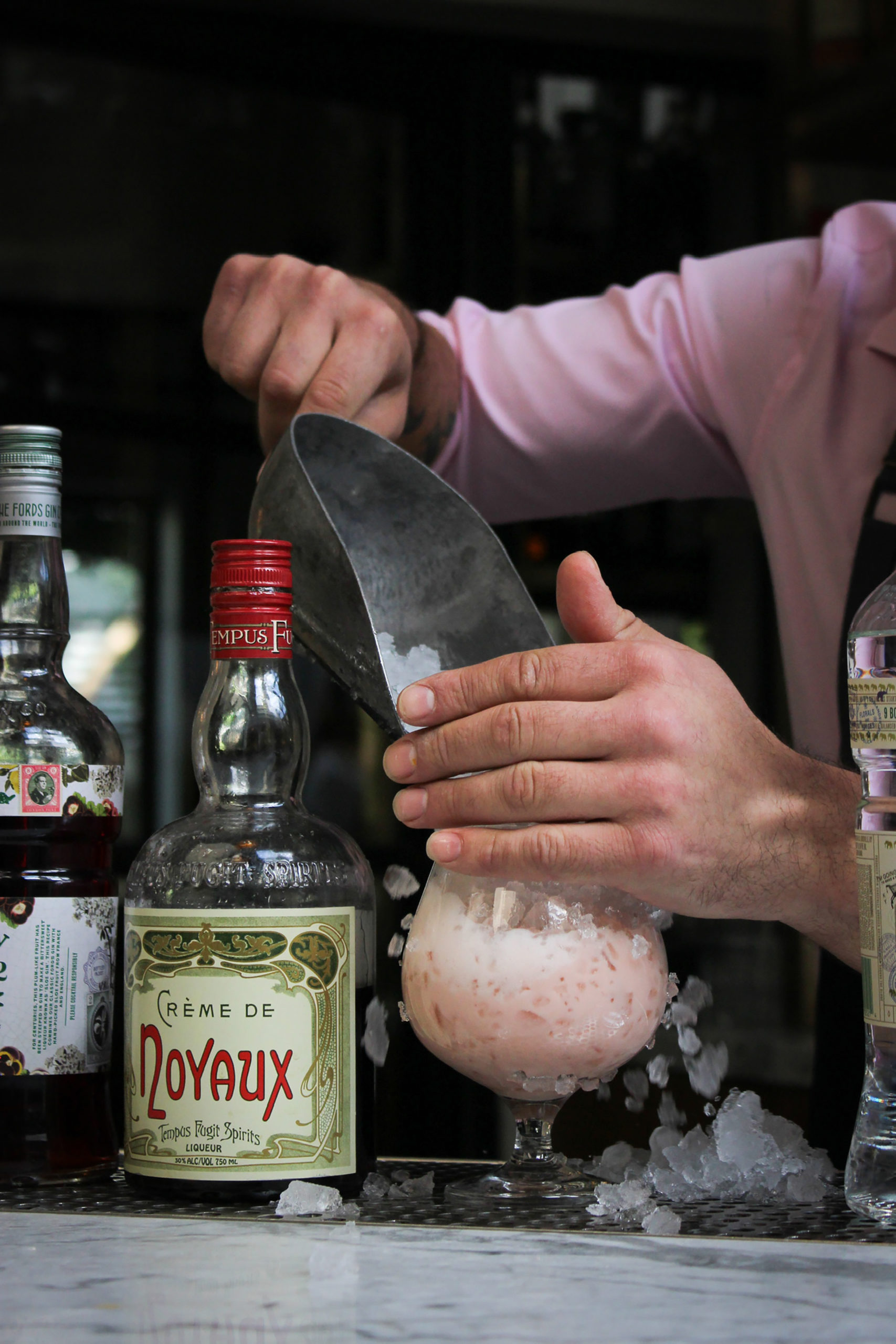 bartender in a pink shirt putting ice in a drink surrounded by bottles